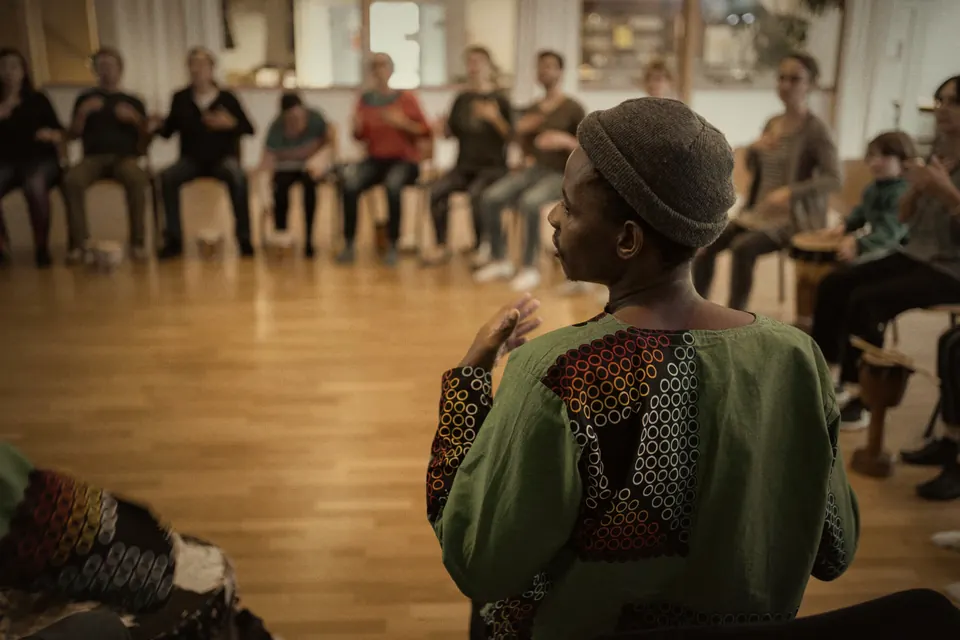 Trommel-Workshop: eine Lehrkraft mit Mütze und grünem Gewand vor einem Stuhlkreis