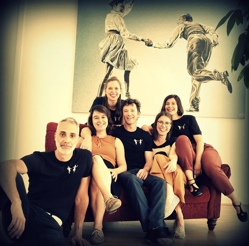 Das Team auf und vor dem roten Sofa im Studio, unter dem Wandbild